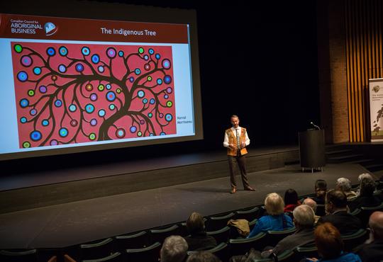 JP Gladu speaks on stage with a photo of Norval Morrisseau's Indigenous Tree on the screen