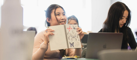 A student looks happy and is holding up their sketchbook which has lots of drawings in it.