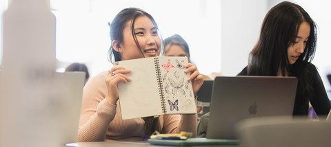 A student at a desk is holding a sketchbook with drawings on the front of it.