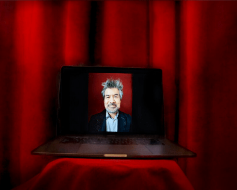 An image of a man in a suit jacket is displayed on a laptop screen with a red curtain background. The laptop in on a red table.