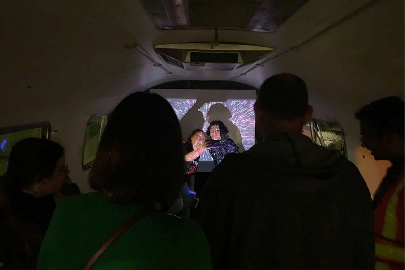 Inside a vintage airstream trailer, two people take a selfie in front of a projected light display.