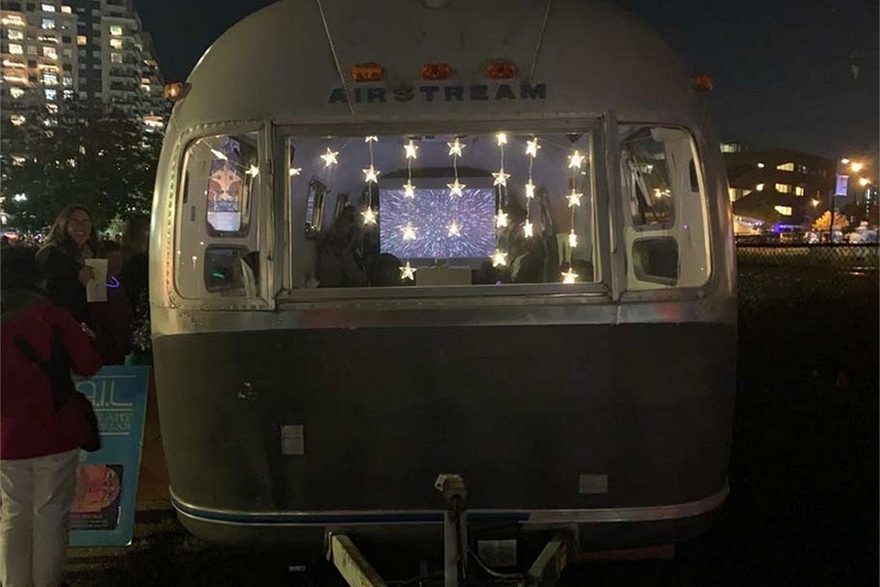 At night in a downtown environment, a vintage airstream trailer, with lighted stars hanging in the window, displays a video of coloured lights.
