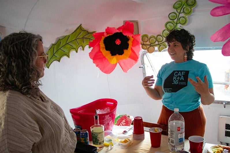 Two people stand at a refreshement table inside a vintage airstream trailer decorated with giant paper flowers.