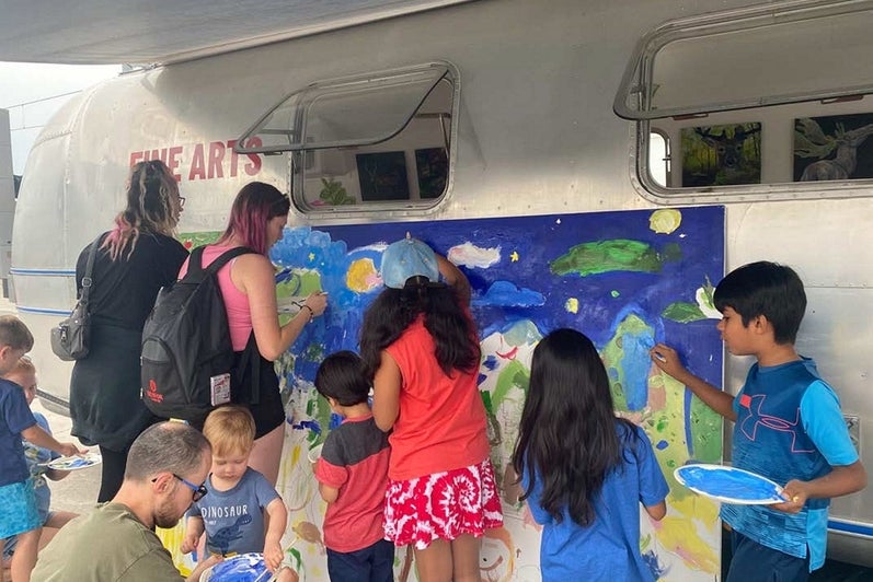 Adult and children paint a landscape on a giant canvas leaning against a vintage airstream trailer.