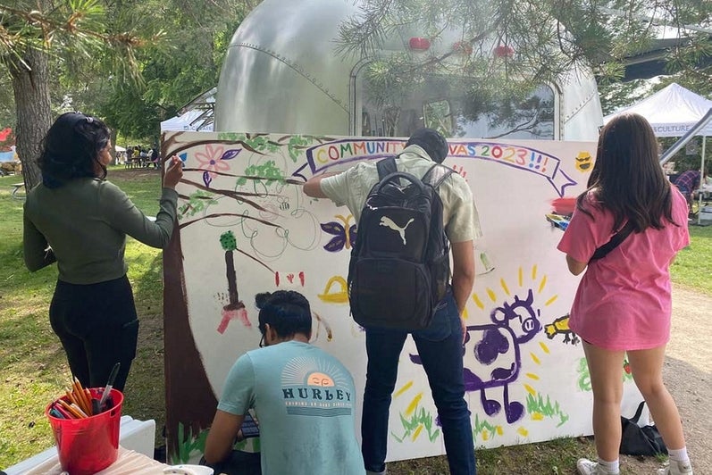 People paint a giant canvas leaning against a vintage airstream trailer. The painting reads "Community Canvas 2023".
