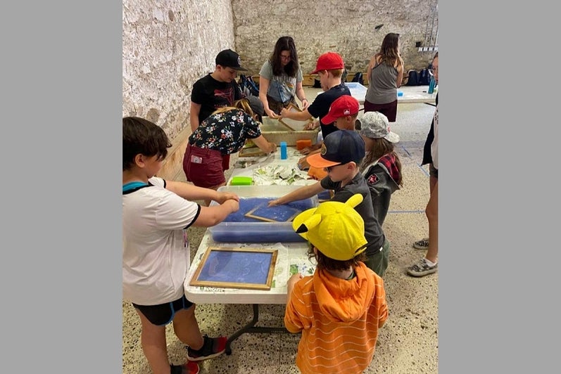 In an old stone building, a group of children gather around table cleaning screens for paper-making.