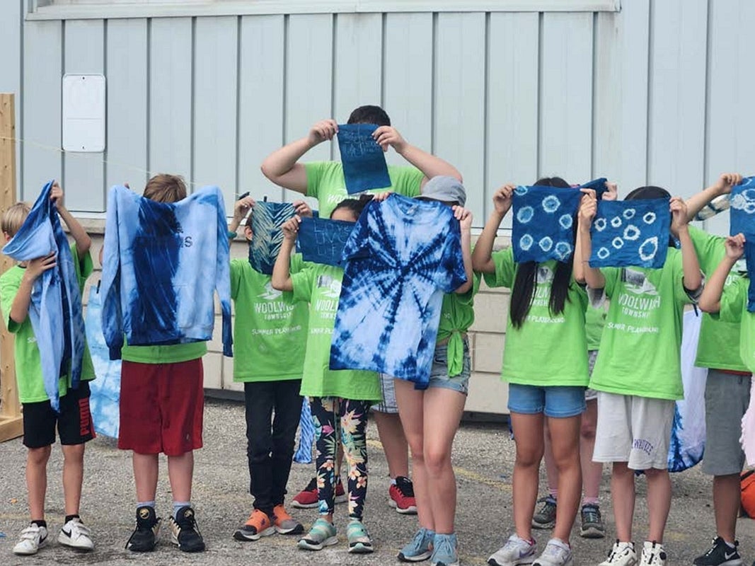 A group of children stand outside a building holding up indigo tie-dyed fabric and clothing