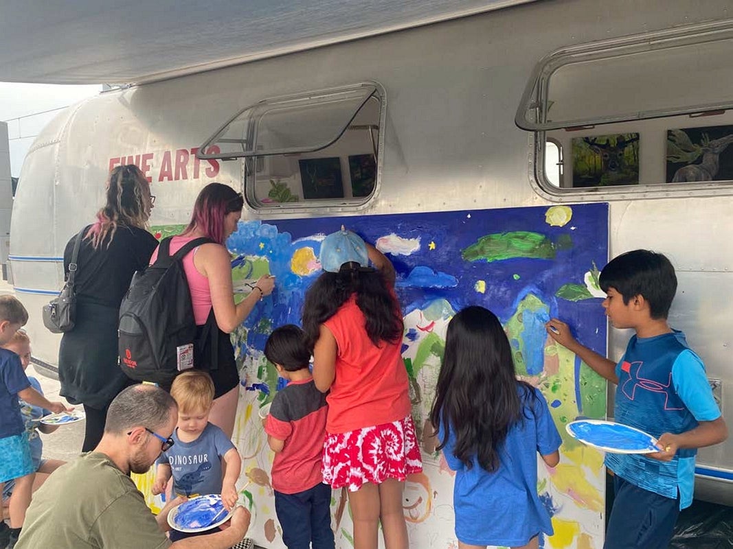 Adult and children paint a landscape on a giant canvas leaning against a vintage airstream trailer.