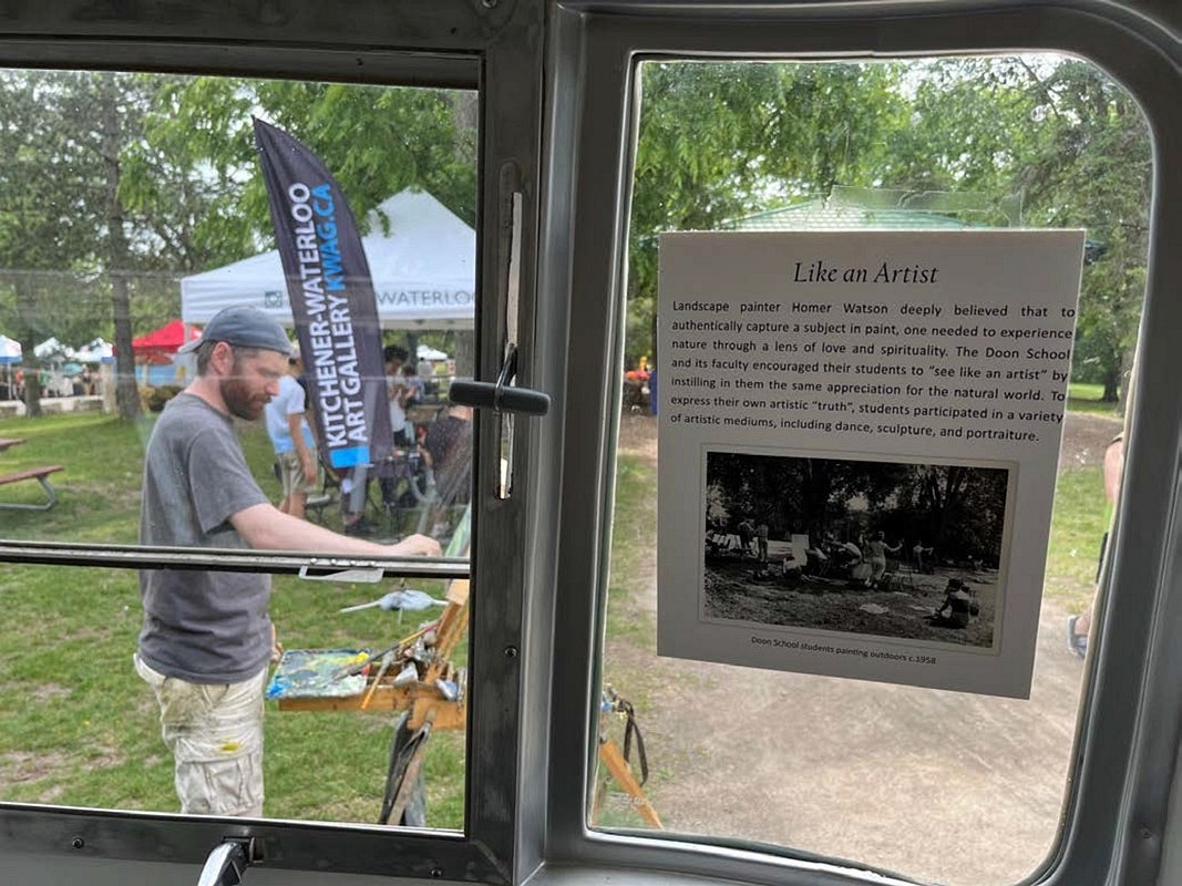 Photographed through the window of a vintage airstream trailer, an artist paints at an easle.  The card in the window explains about an artist experiencing nature.