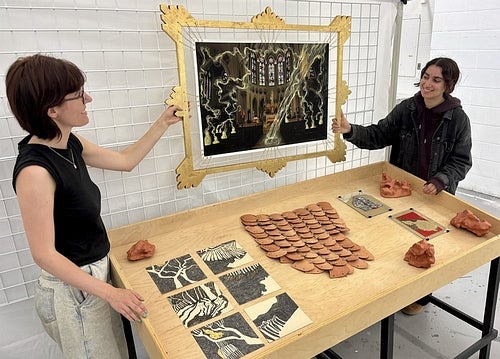 Two people hold up a dark photograph in the gilt frame with a table in front of them containing multiple small art prints and clay sculptures.