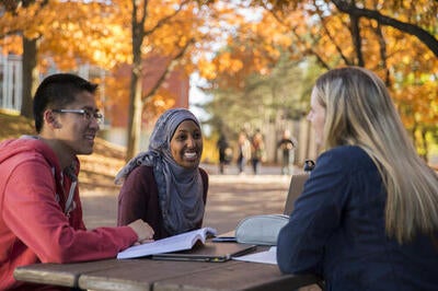 Three students facing each other as they sit on a picnic bench on campus and talk.