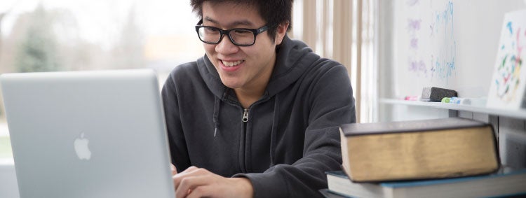 a student sits at a desk, smiling at a computer with a stack of textbooks in the foreground