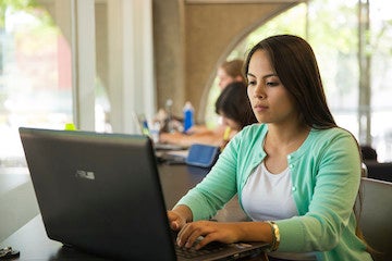 A student sitting at a desk inside a building on campus and working on a laptop.