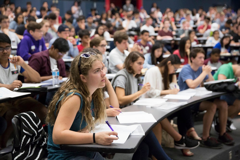 A group of students in a lecture hall on campus listening to a professor and taking notes.