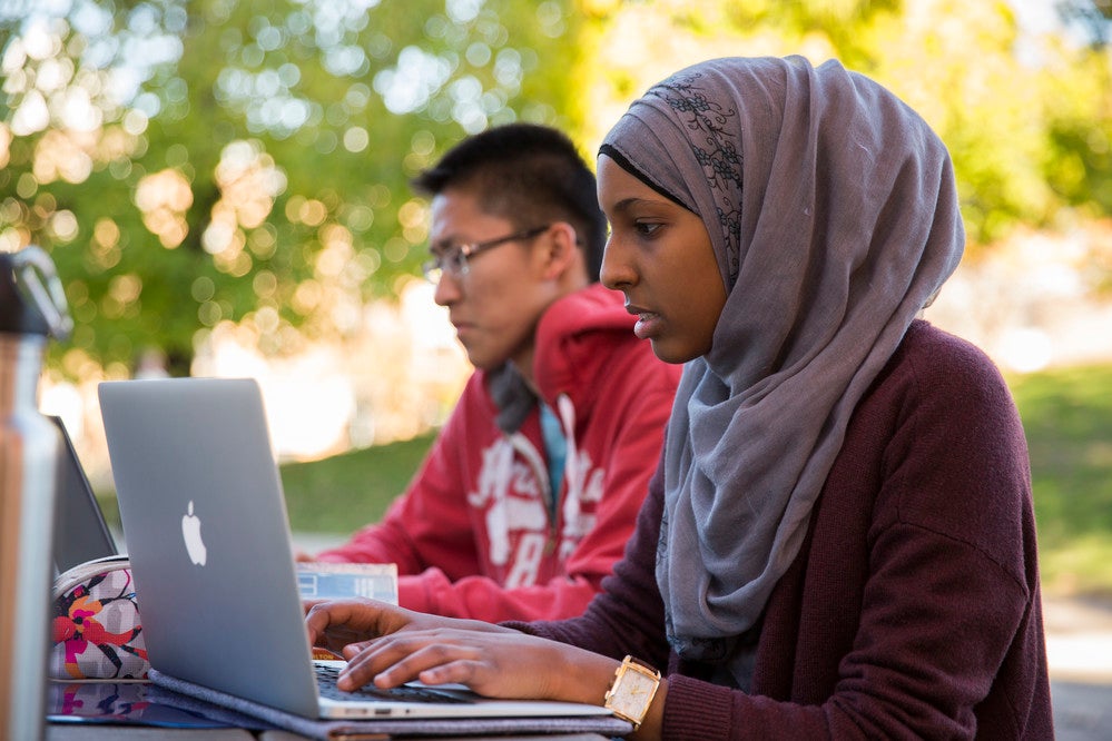 Two students sitting outside on a picnic bench. Both are looking at their own laptops.