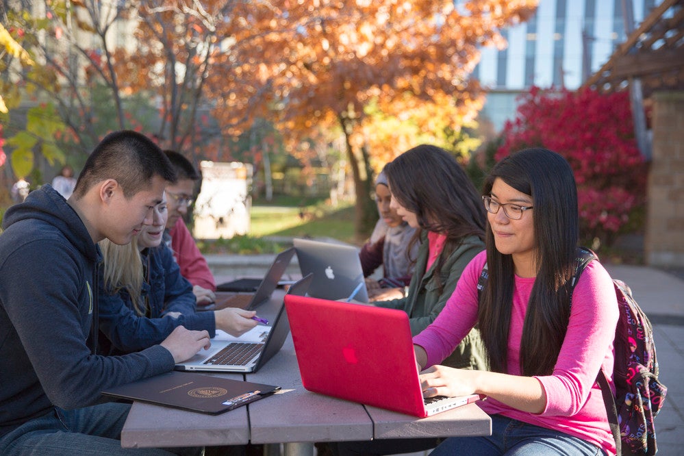 Four students sitting at a picnic bench on campus working on their laptops.