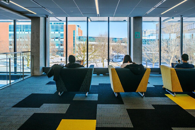 Three students sitting in chairs in a building on campus, looking outside through windows