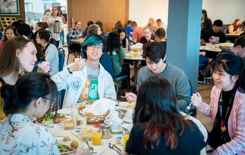 A group of students sitting in the Conrad Grebel cafeteria. You have the group of 6 students in the fore ground, and then other groups of students sitting in the background. All are eating a meal.