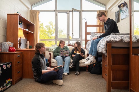 A group of 4 students sitting in a Conrad Grebel room. 1 student is sitting on the floor agains the dresser, two students are sitting on a couch, and one student is sitting on a raised bed. 