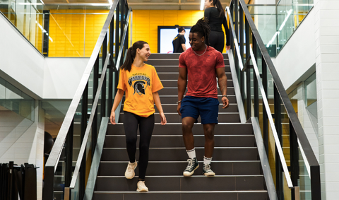 Two students walking down a set of stairs in athletic gear inside the PAC.
