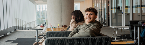 A student smiling at the camera while sitting in a lounge chair inside a modern campus building, with another student beside them working on a laptop near large windows and study tables.