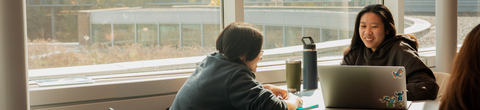 Two girls sitting across from each other and sitting infront of their computers smiling