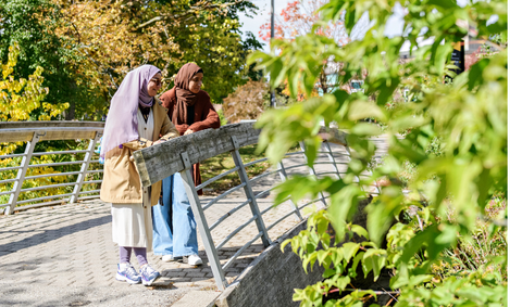 two students standing on the bridge between main campus and health services building overlooking the river