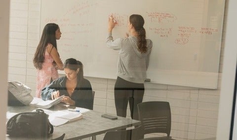 2 students working out a problem on a whiteboard while another studies