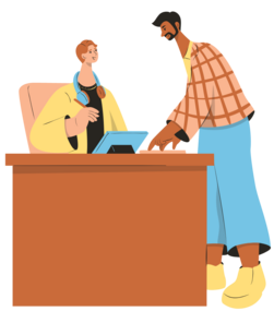 A student sitting at a desk. Another older looking student approaches the desk and is talking to the first student.