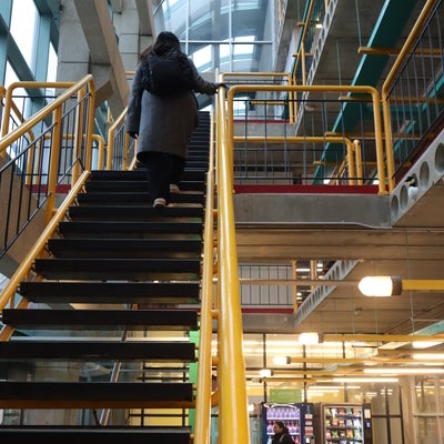 Person walking up a staircase with yellow railings inside the University of Waterloo Davis Center Building, with vending machines and structural beams visible below.
