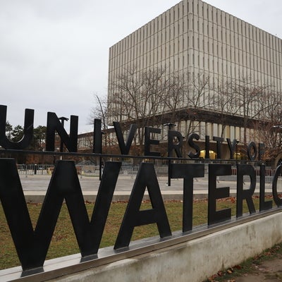 Large black ‘University of Waterloo’ sign displayed outdoors on campus, with the Dana Porter Library building in the background on an overcast day.