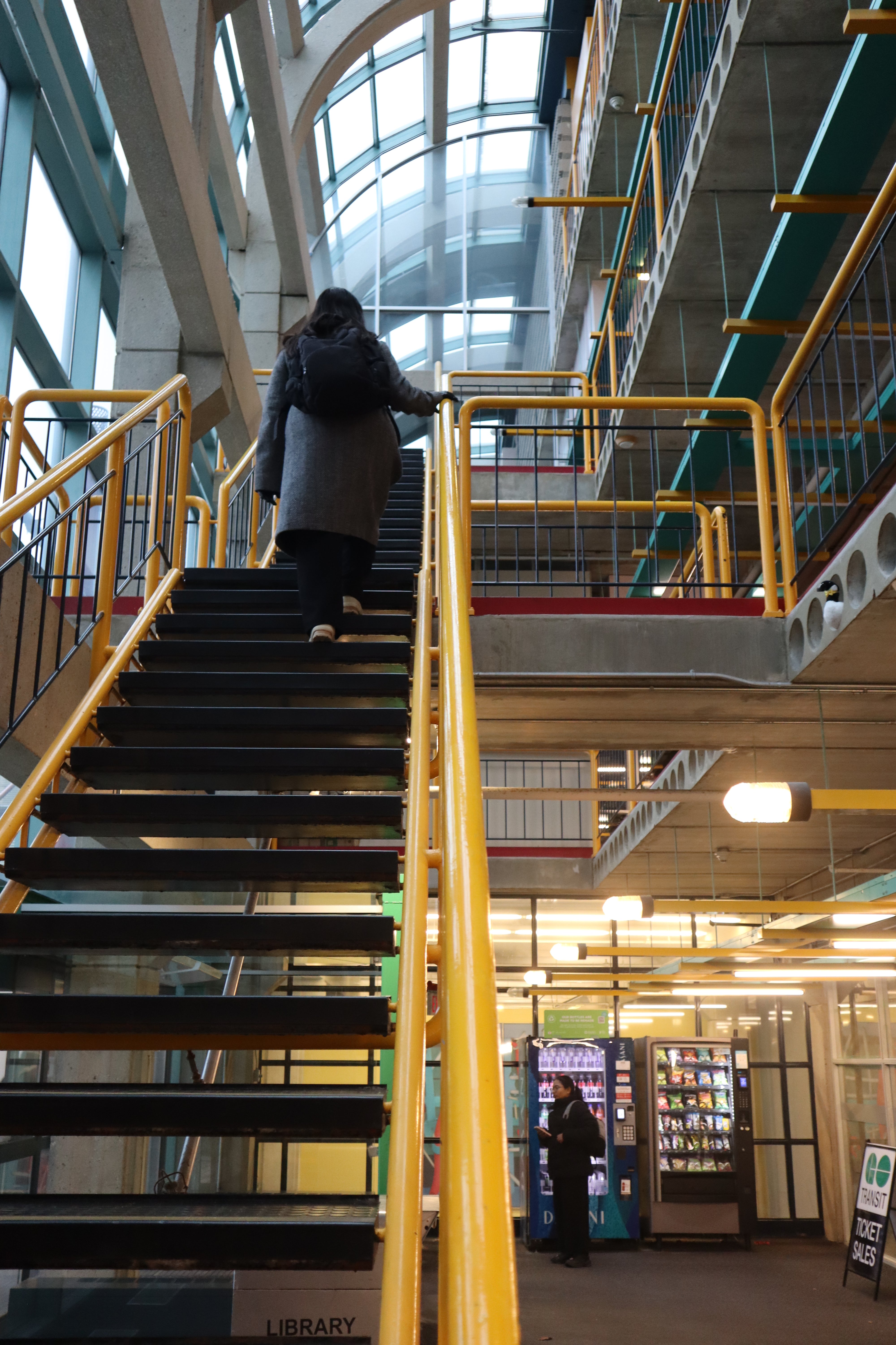 Person walking up a staircase with yellow railings inside the University of Waterloo Davis Center Building, with vending machines and structural beams visible below.