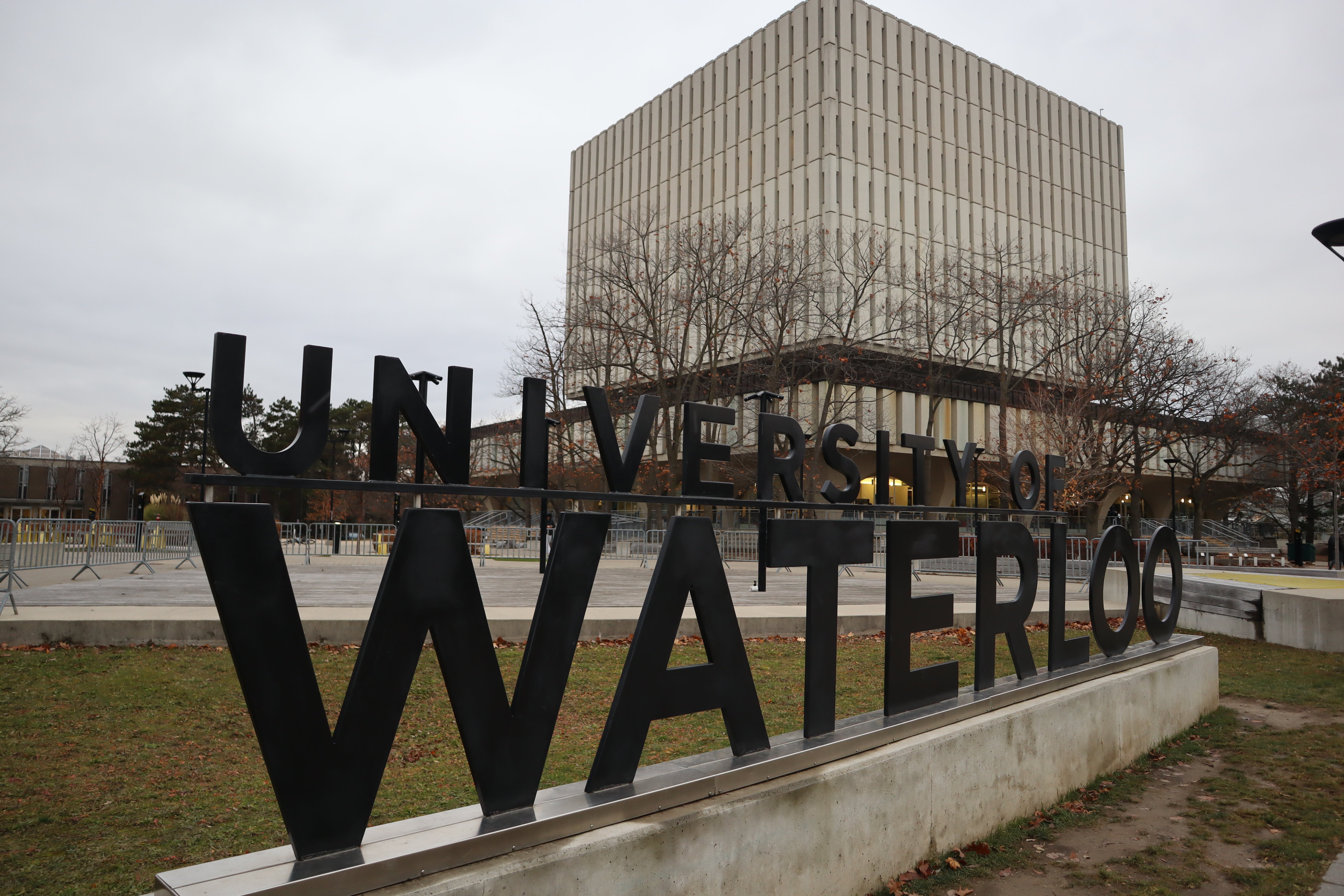 Large black ‘University of Waterloo’ sign displayed outdoors on campus, with the Dana Porter Library building in the background on an overcast day.