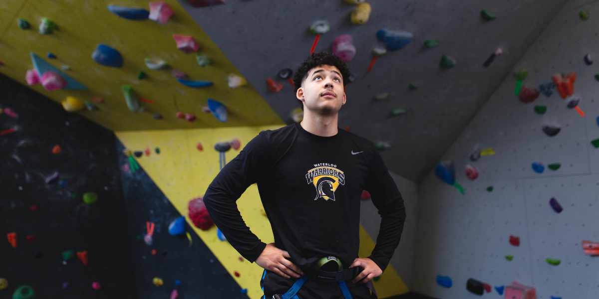 Young man in front of rock climbing wall, with hands on hips, looking up