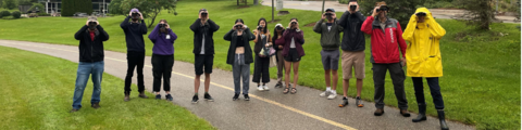 11 people stand outside holding binoculars up to their eyes.