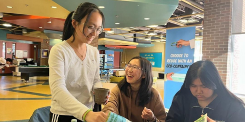 3 people stand around a booth. One is laughing, one is eating, and one is focused on the table.