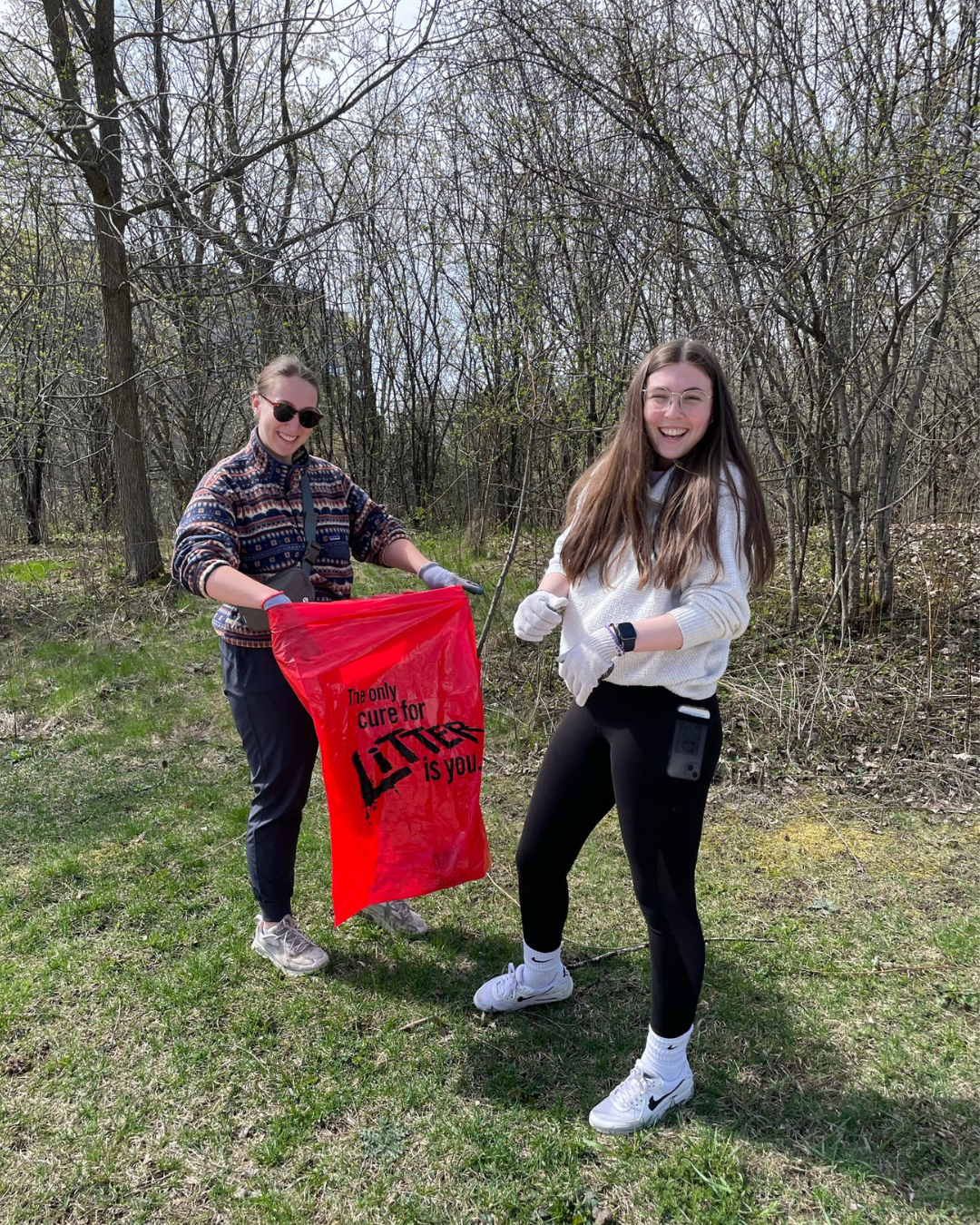 Two students stand in front of a woodlot with a red garbage bag. 