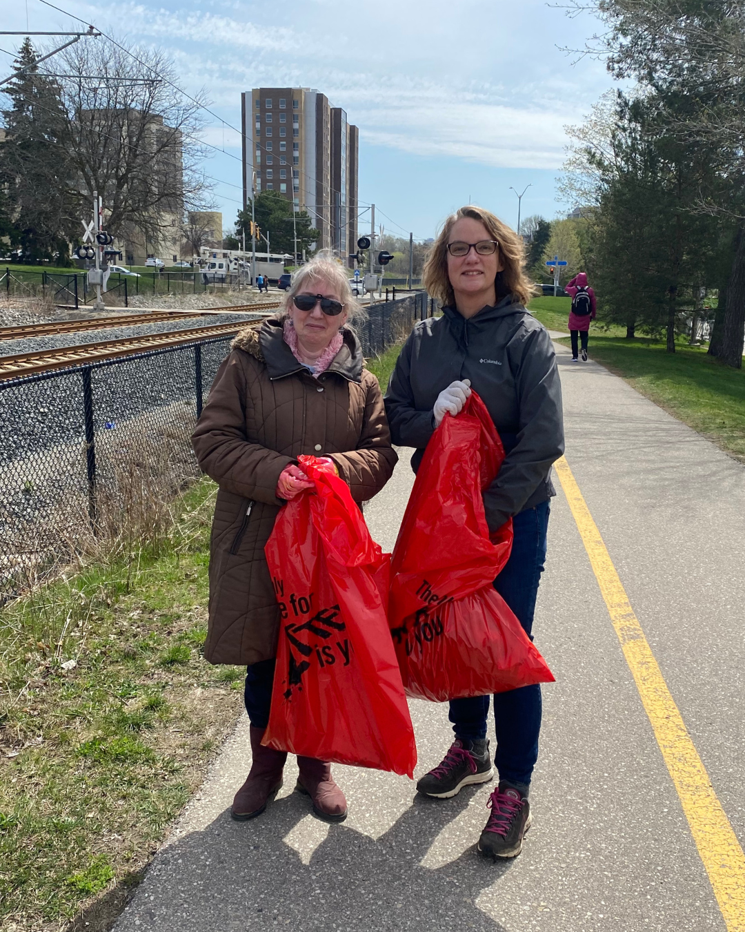 Two staff volunteers stand on the trail by the ION tracks with red garbage bags. 