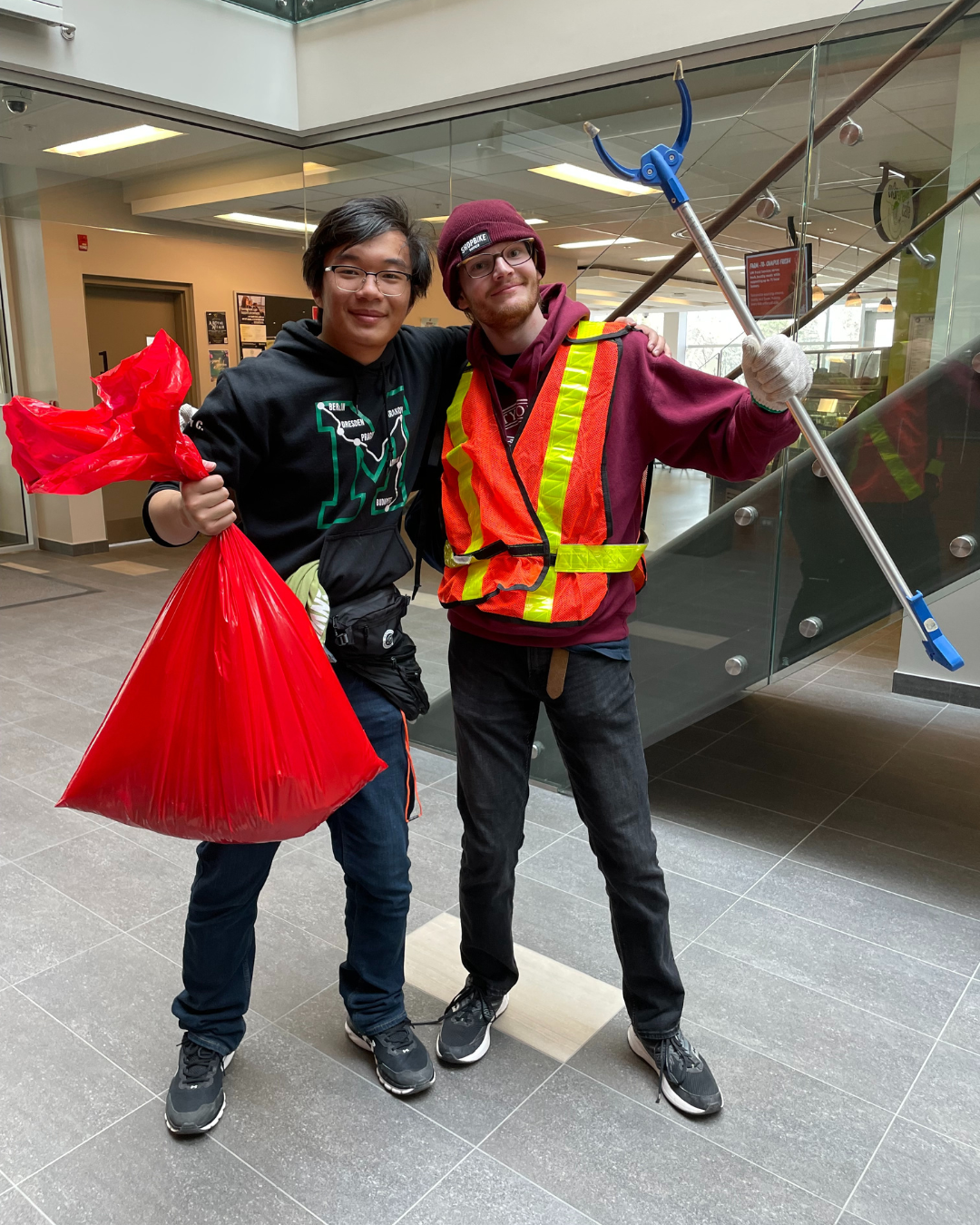 Two student volunteers (one wearing a safety vest) stand in the EV3 atrium with a garbage picker and a red garbage bag. 