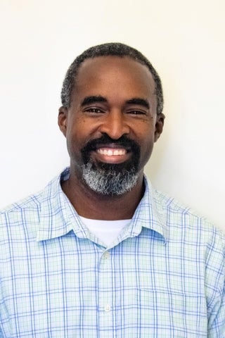 Photograph of man with a black beard smiling at the camera. Wearing a light blue checkered dress shirt and white undershirt underneath. 