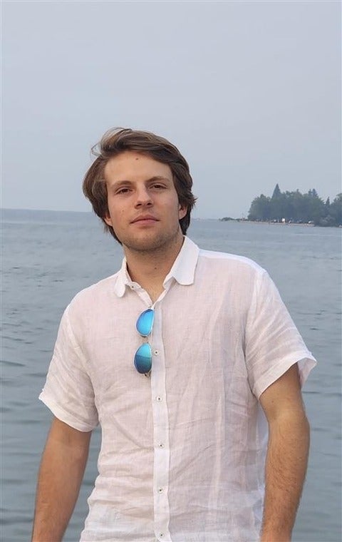 Photograph of a young man with mid-length brown hair smiling at the camera. He is wearing a white shirt and has sunglasses tucked into his collar. 