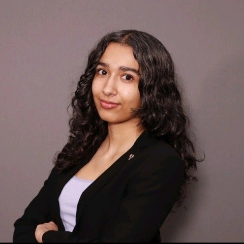 Picture of a woman with long dark brown coloured hair, smiling at the camera, with her arms folded across her chest. She is wearing a black blazer with white blouse.
