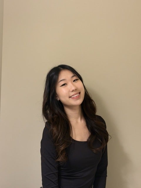 Photo of woman smiling against the backdrop of a taupe coloured wall. The woman photographed is wearing a black long-sleeved shirt, and hair is dark brown/black with loose curls in them. 