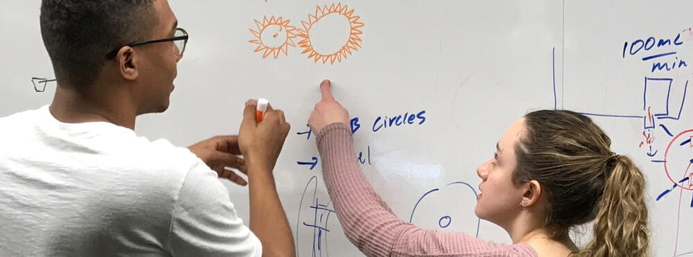 Two students (one male, one female) collaborate at a large whiteboard
