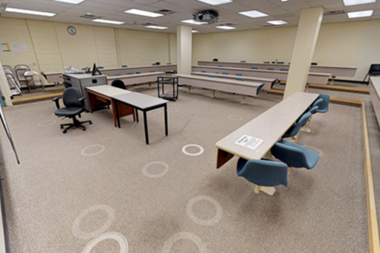 Classroom space with fixed tables and seating configured in a horseshoe around a front dais. Pillars block some visibility.