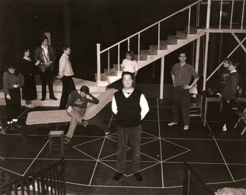 Man in a sweater vest standing with arms behind his back, surrounded by a group of people