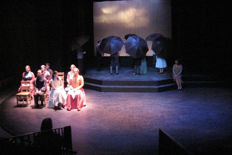 Group of people sitting on stage with another group in the background holding umbrellas