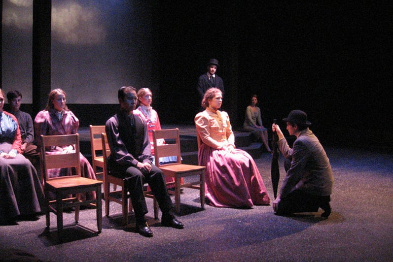 Group of people sitting on stage facing a man kneeling with an umbrella