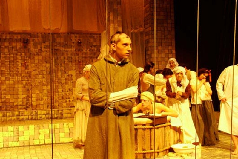 Man with a shaved head in the foreground with other cast of Marat/Sade performing on stage