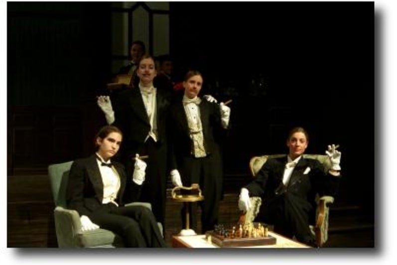 Cast of The Club dressed in tuxedos lounging around a chess board